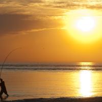 En fisker står i vandkanten ved Vesterhavet. Der er en smuk, orange solnedgang i baggrunden