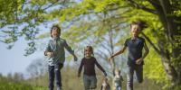 Tre børn i blåpt tøj løber på markvej med grønne bøgetræer i baggrunden
